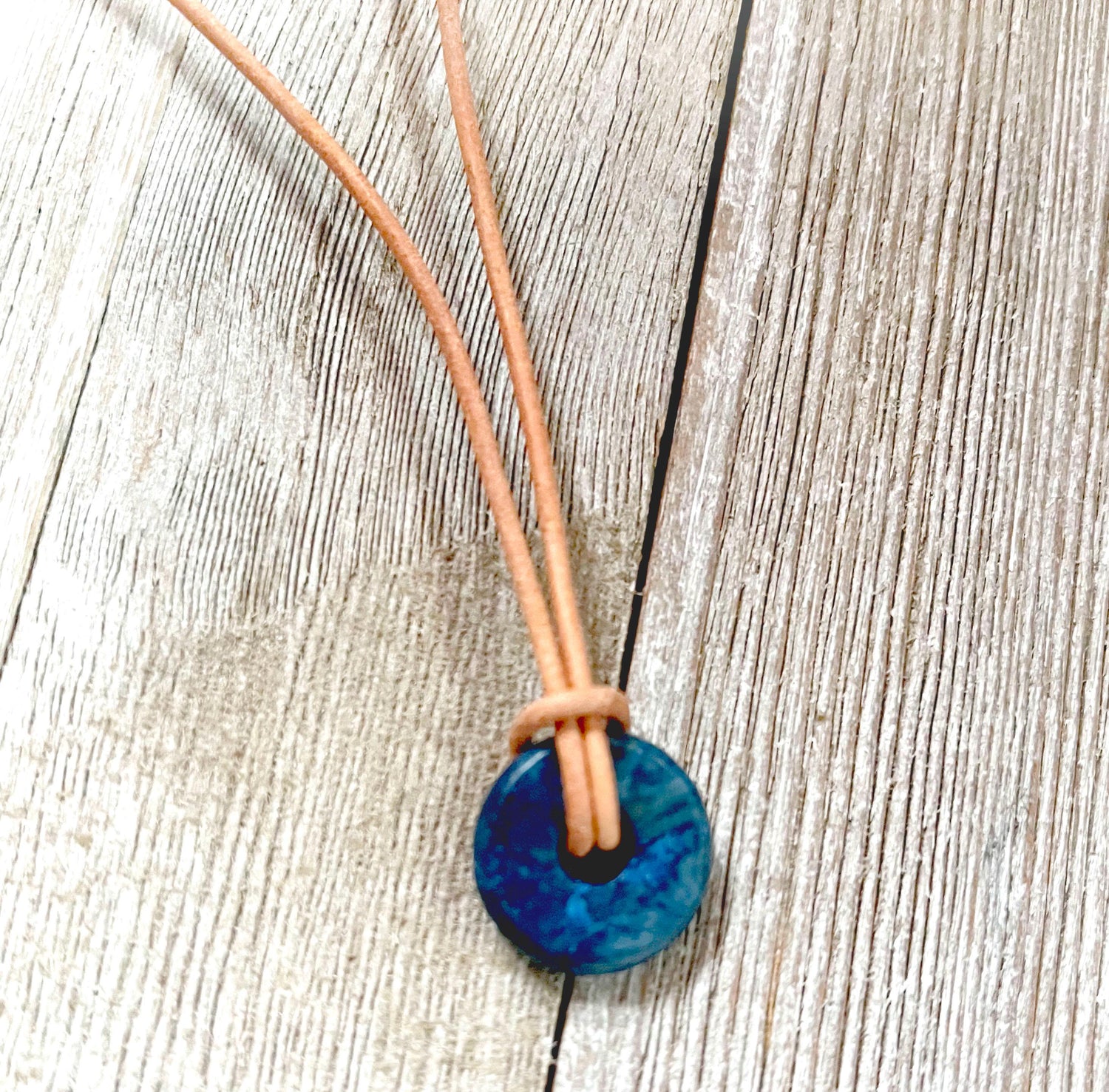 The Minimalist Lapis Lazuli Necklace features a blue circle stone pendant on an Italian leather cord, tied in a simple knot and displayed on a weathered wooden surface.