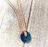 The Minimalist Lapis Lazuli Necklace features a blue circle stone pendant on an Italian leather cord, tied in a simple knot and displayed on a weathered wooden surface.