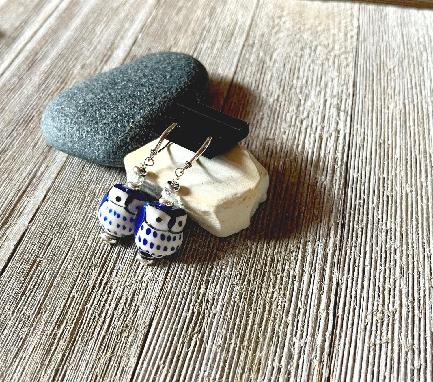 Handcrafted Glass Owl Earrings with color change topper beads and blue-white patterns are displayed on stacked stones atop a rustic wooden surface.