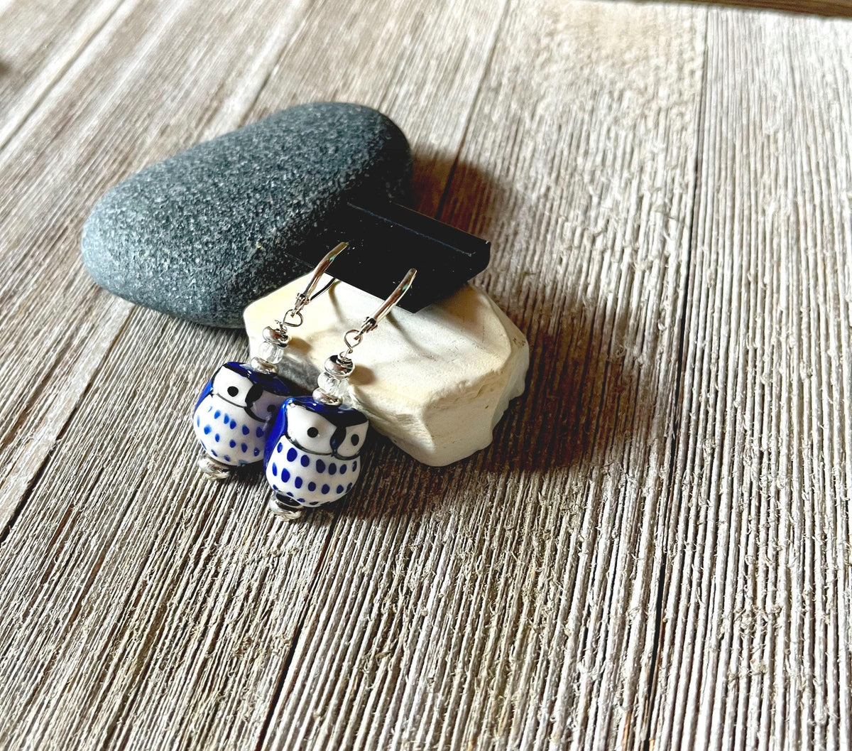 Handcrafted Glass Owl Earrings with color change topper beads and blue-white patterns are displayed on stacked stones atop a rustic wooden surface.