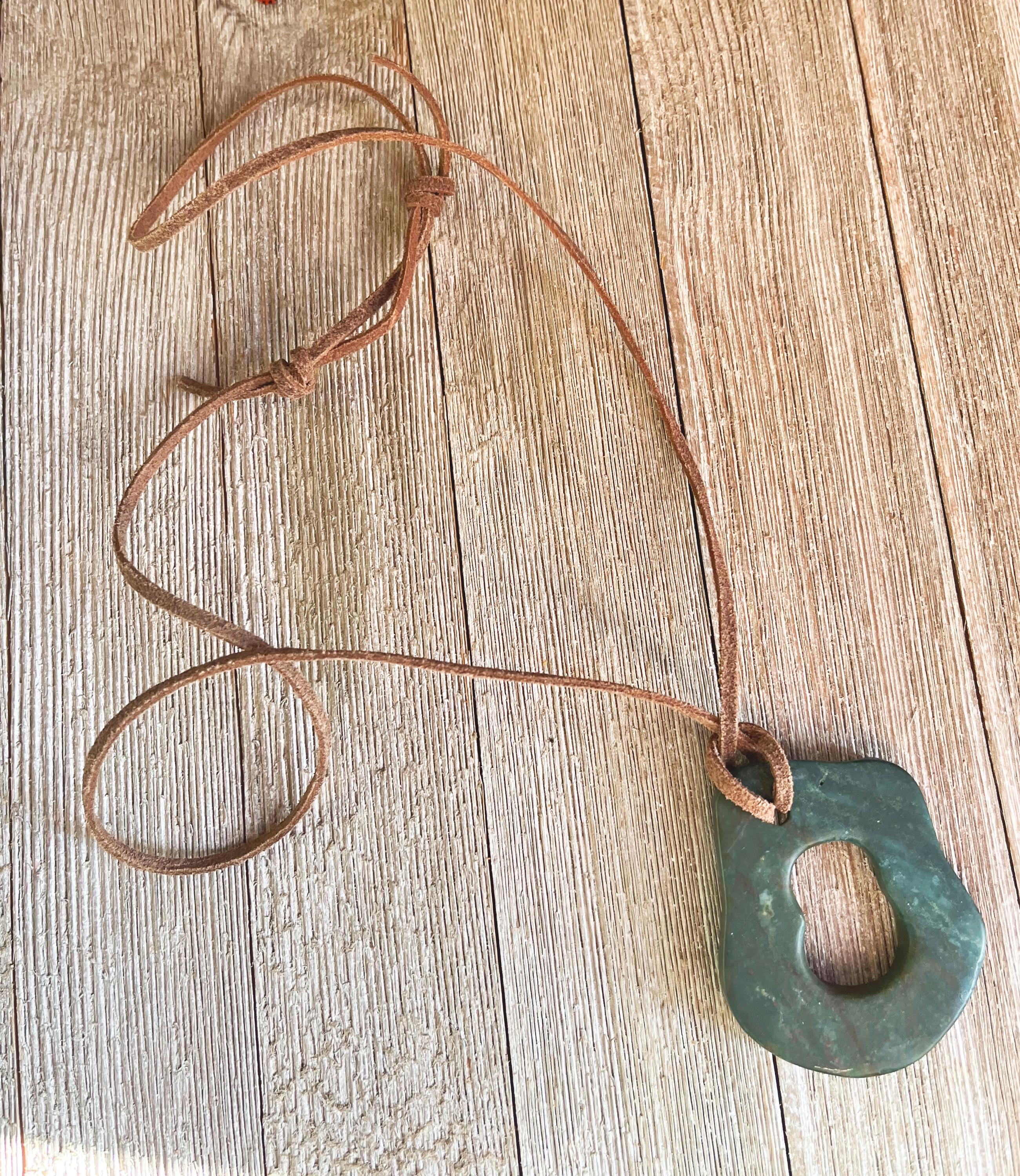 The Handcrafted Green Aventurine Pendant Necklace features an irregular greenstone pendant with a central hole on an adjustable leather cord, displayed on a light wooden surface.