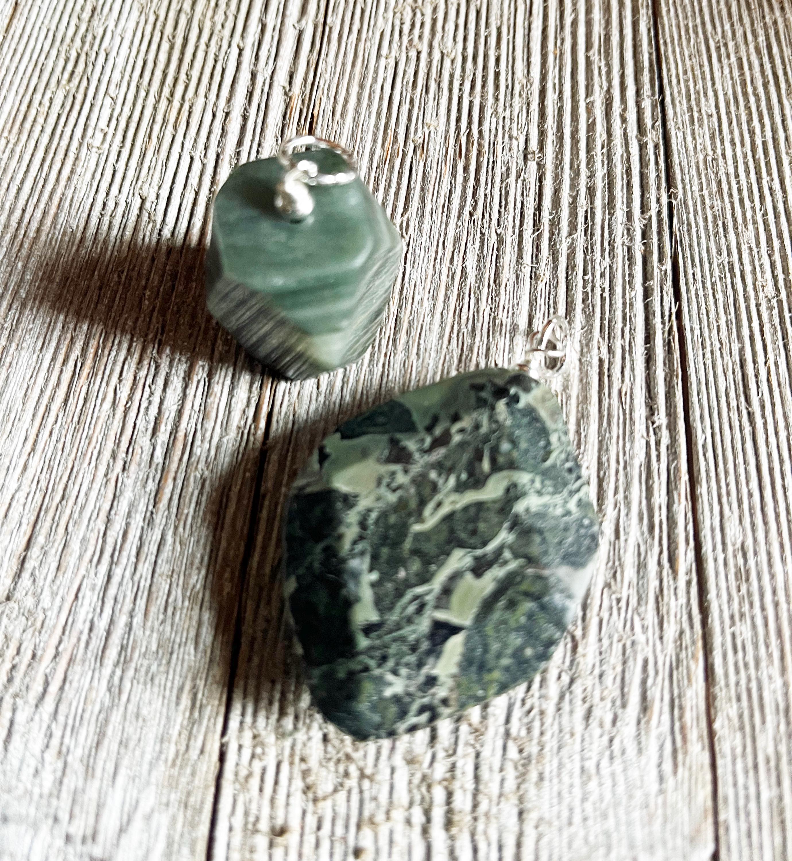 The Chunky Style Masc Forest Green Stone Pendant—an earthy gemstone on a 5mm cord with a silver bail—rests on rustic, light wood, showcasing its forest green color and marbled finish for an eye-catching natural necklace.
