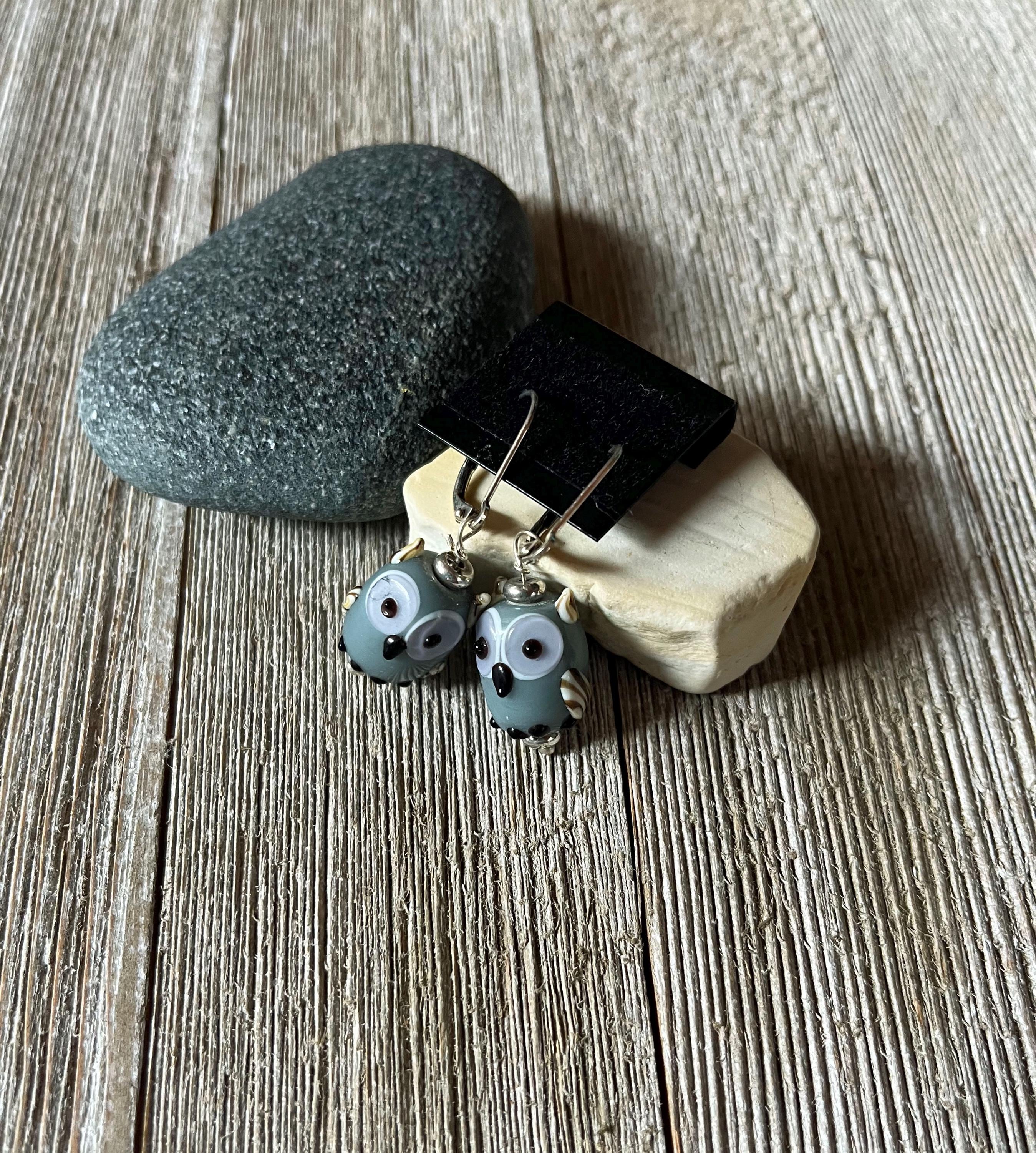 Enchanting Handcrafted Animal-Themed Owl Earrings with blue and white details are displayed on a black stand atop smooth stones—unique whimsical woodland jewelry, perfect as a bird lover gift.