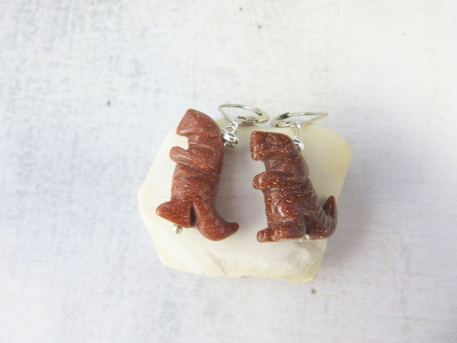 Goldstone T-Rex Dinosaur Earrings—handcrafted with shimmering brown dinosaur charms and sterling silver lever backs—make a striking animal jewelry gift, as shown on a white stone background.