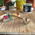 A bundle of sage with colorful strings and beads rests next to the Zodiac Sign Coffee Scoop – Beaded Zen Herb Spoon, Fire Element, on a wooden surface. Behind them are potted plants and a mug featuring a hamsa hand design.