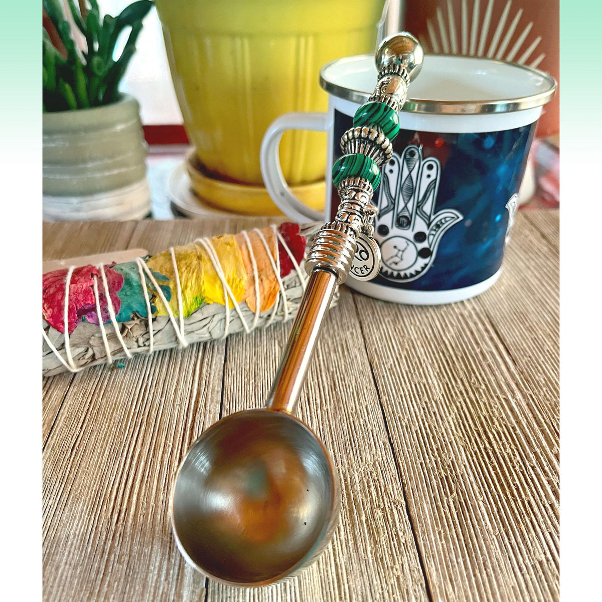 The Zodiac Water Element Spoon—Beaded Zen Coffee Scoop for Pisces, Cancer, and Scorpio—rests on wood beside a vibrant sage bundle and a hamsa mug, with a yellow pot and other items blurred in the background.
