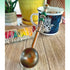The Zodiac Water Element Spoon—Beaded Zen Coffee Scoop for Pisces, Cancer, and Scorpio—rests on wood beside a vibrant sage bundle and a hamsa mug, with a yellow pot and other items blurred in the background.
