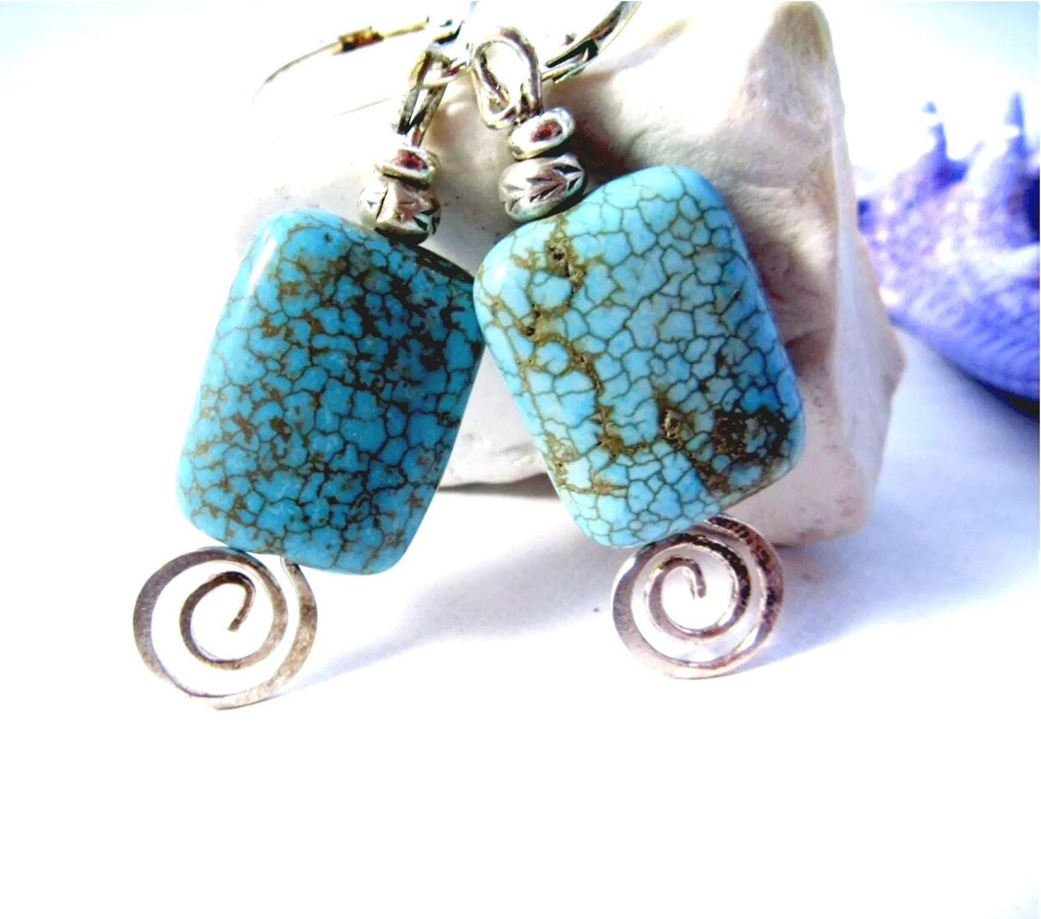 Turquoise Howlite Earrings: Thai Silver Swirl Dangle feature rectangular blue stones with dark veins, silver beads, and spiral charms. Artisan designed jewelry, displayed on a white and light purple background.