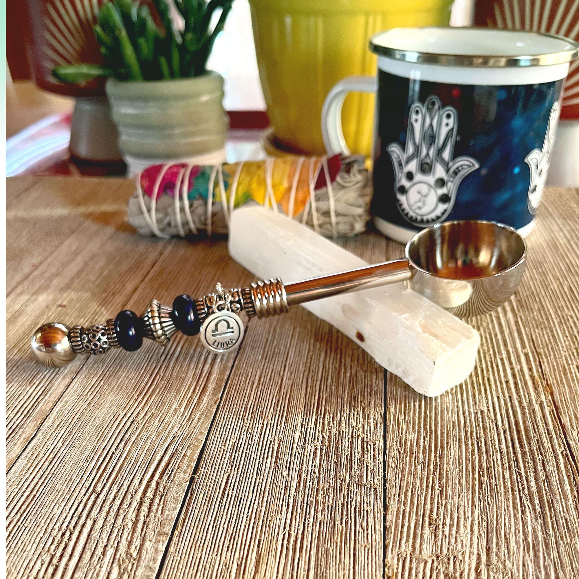 The Zodiac Herb Scoop with Beaded Handle: Zen Coffee Spoon rests on a white crystal stick atop a wooden table, with a Hamsa mug, yellow plant pot, and wrapped sage bundle visible in the background.