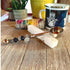 The Zodiac Herb Scoop with Beaded Handle: Zen Coffee Spoon rests on a white crystal stick atop a wooden table, with a Hamsa mug, yellow plant pot, and wrapped sage bundle visible in the background.