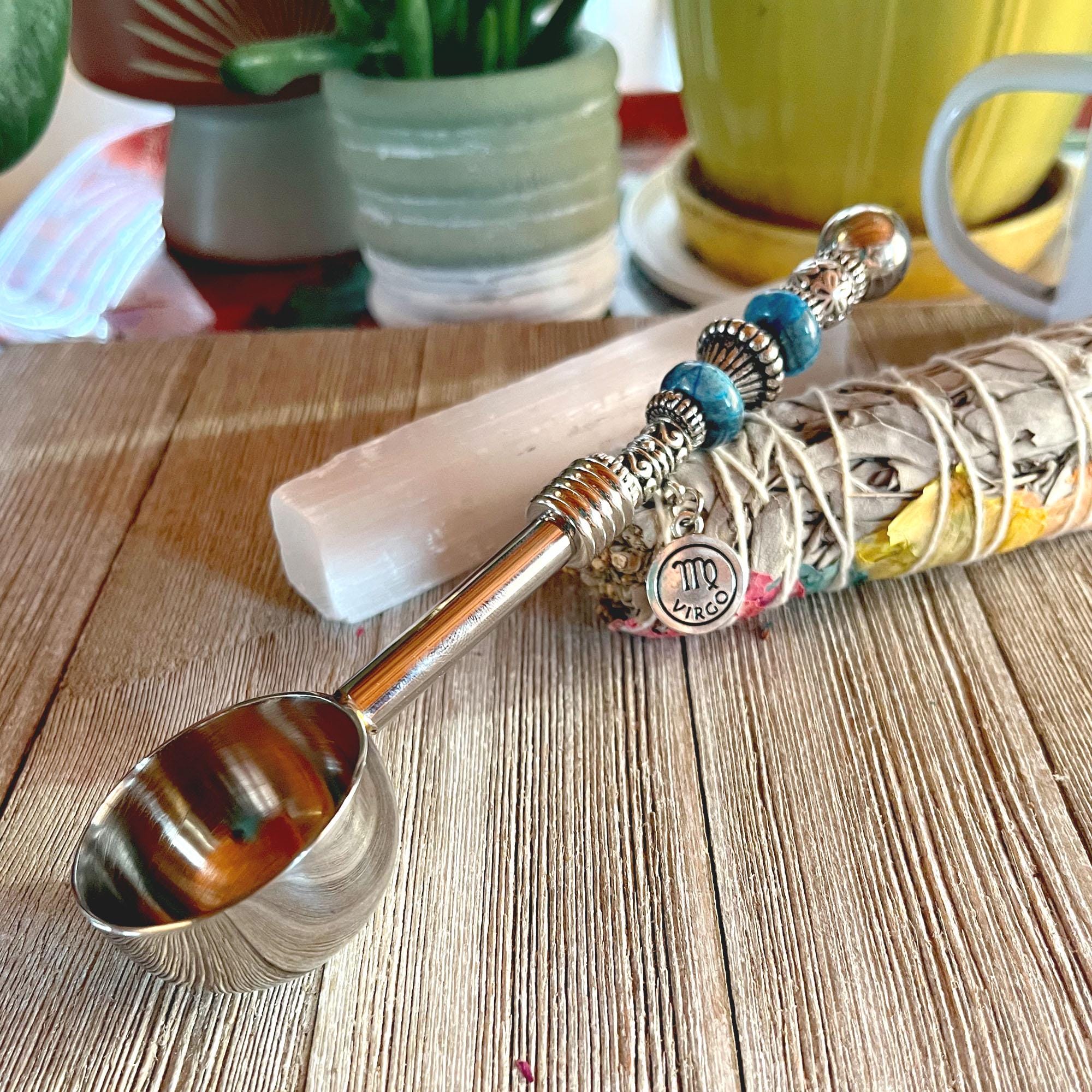 The Zodiac Coffee Scoop – Beaded Handle, Zen Herb Spoon for Earth signs (Virgo, Capricorn, Taurus) rests on a wooden surface with sage, selenite crystal, plants, and a yellow mug in the background.