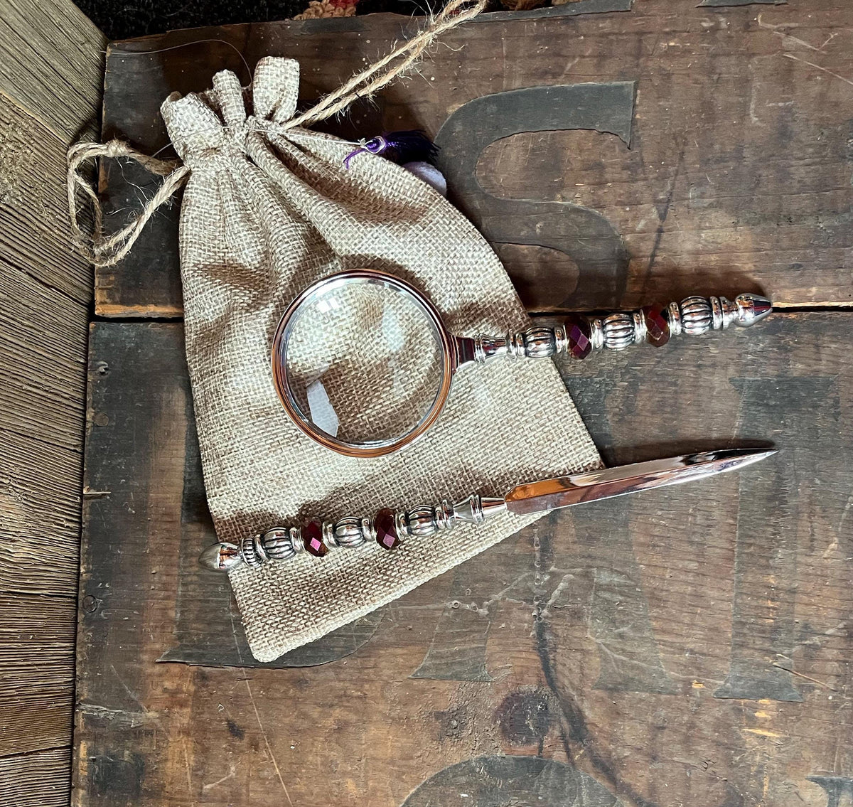 The Purple Magnifier and Letter Opener - Beaded Desk Set features ornate silver and red handles, displayed on a burlap pouch atop a rustic wooden surface.