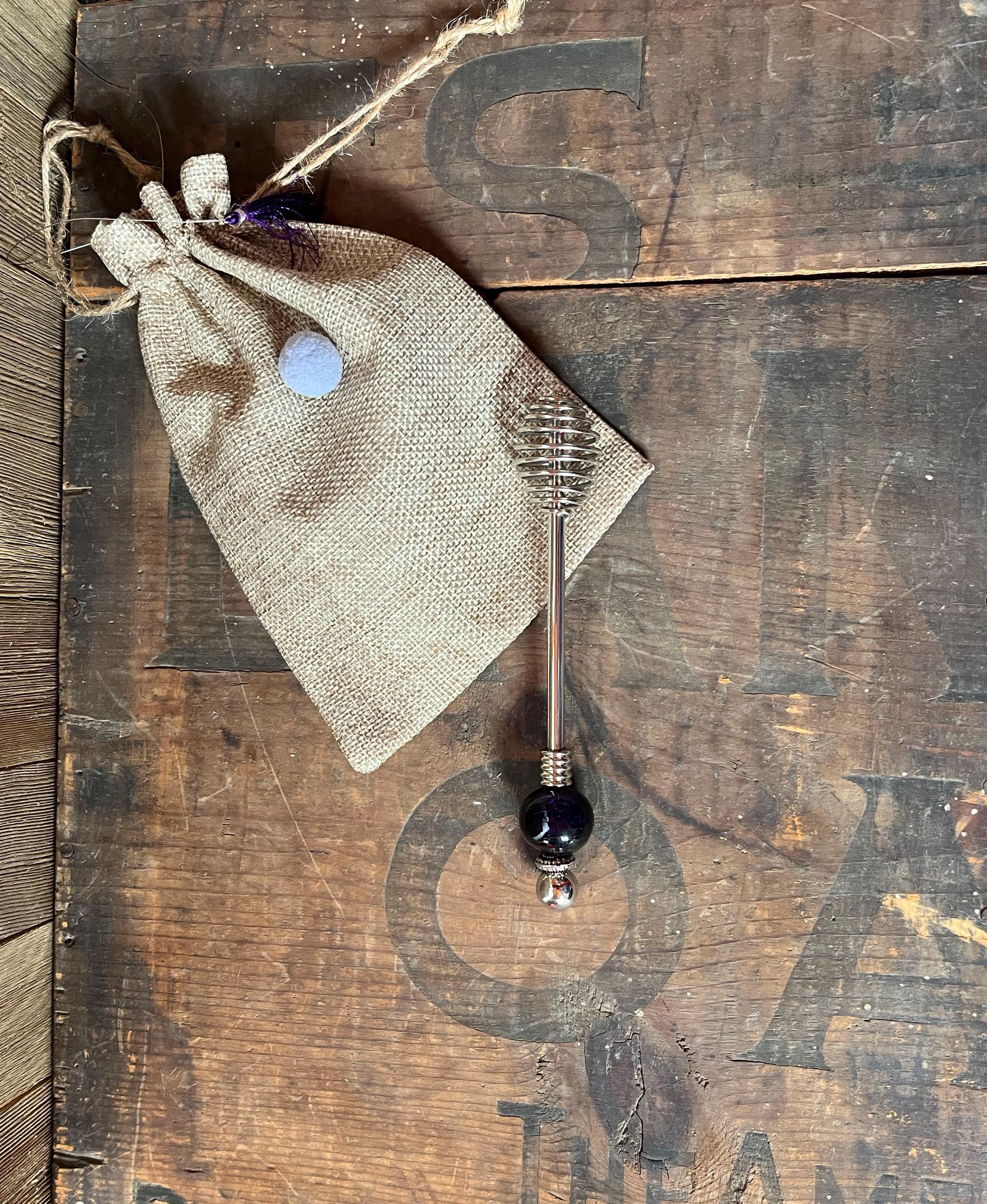 A purple glass honey drizzler with an artisan bead accent rests on a rustic wooden surface beside a burlap drawstring pouch—an ideal kitchen gift.