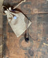 A purple glass honey drizzler with an artisan bead accent rests on a rustic wooden surface beside a burlap drawstring pouch—an ideal kitchen gift.