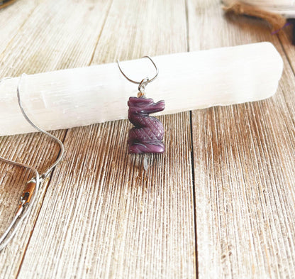 The Purple Dragon Pendant Necklace: Fantasy Glass Jewelry features a spiral-carved purple stone on a silver chain, displayed atop a white crystal wand on a rustic wooden surface.