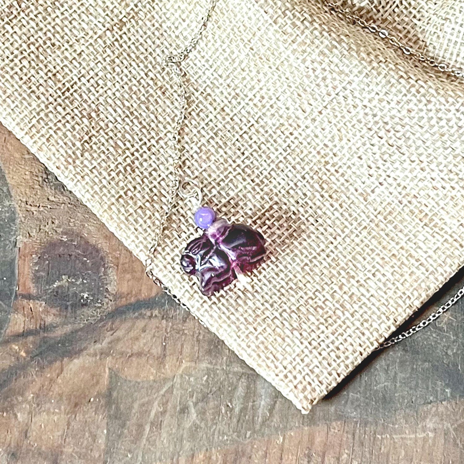 The Bunny Rabbit Necklace features a playful hare pendant and a round amethyst-like lavender bead, beautifully displayed on burlap fabric over rustic wood—an ideal jewelry gift for amethyst lovers.