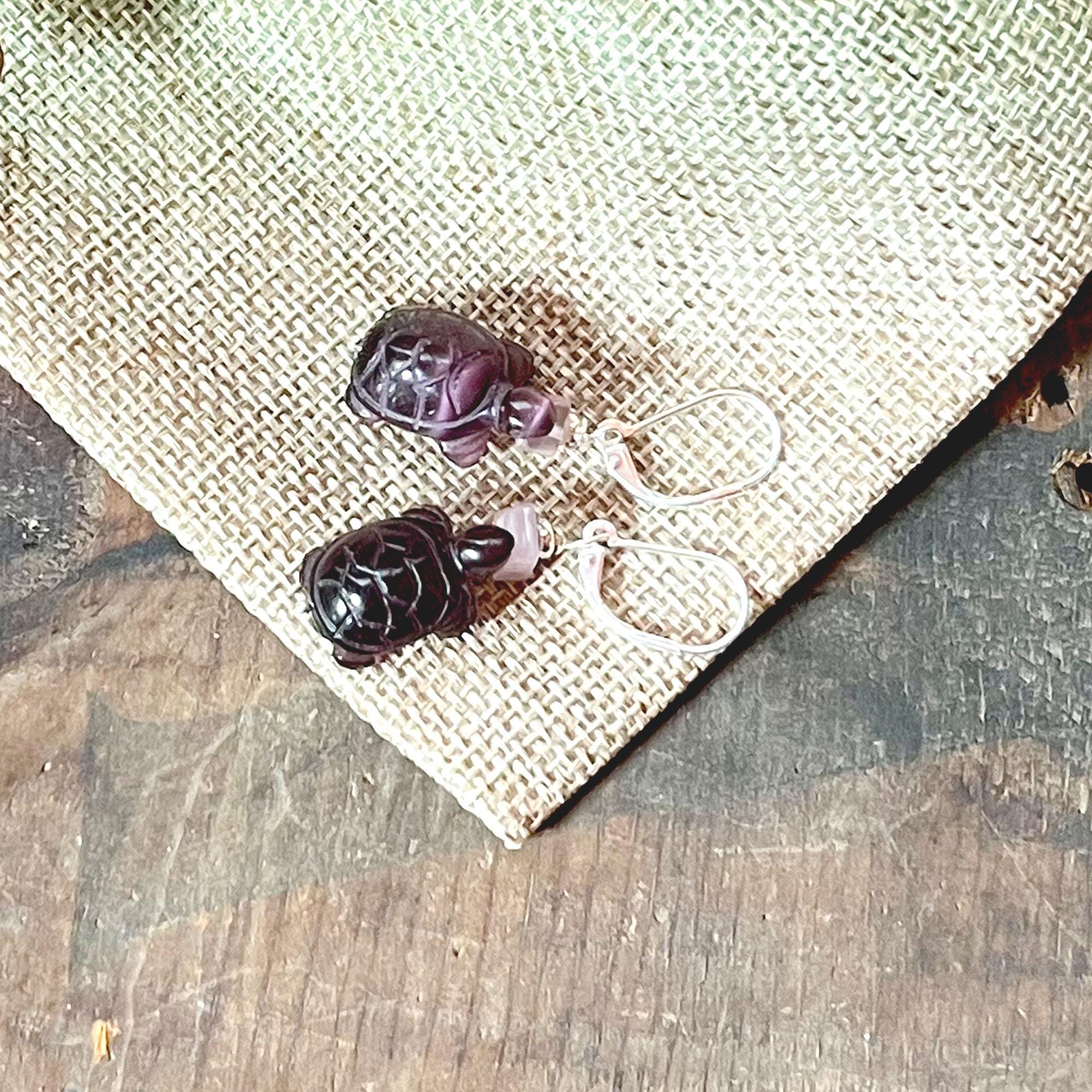 Tortoise Earrings: Optic Amethyst Purple Glass Animal Jewelry with silver hooks, displayed on textured beige fabric atop rustic wood—ideal for animal jewelry lovers.