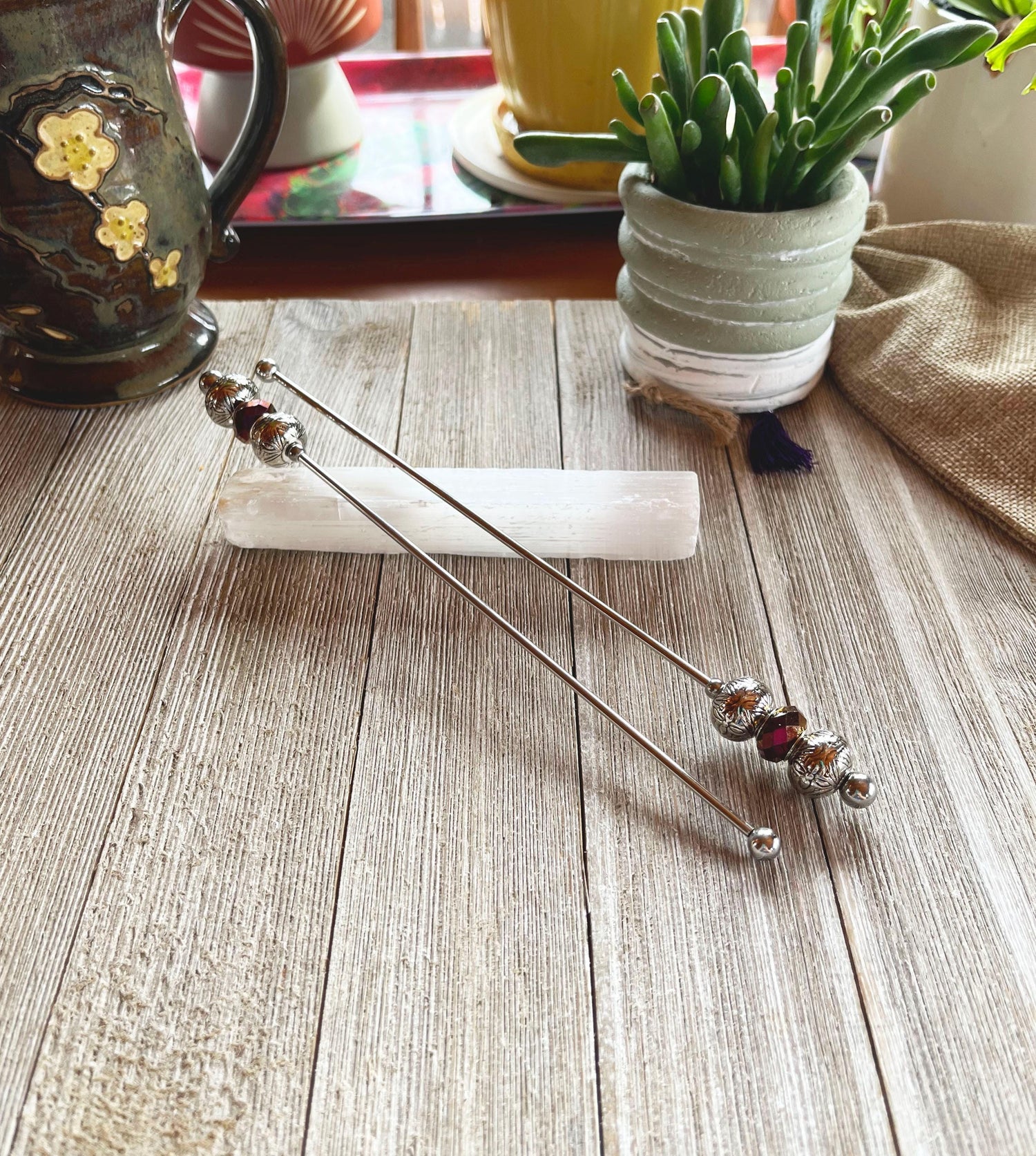 Two Purple Faceted Glass Beaded Drink Stir Sticks—elegant cocktail decor—rest on a white crystal atop wood. In the background are a ceramic cup, potted plant, and other decor. Set of 2.