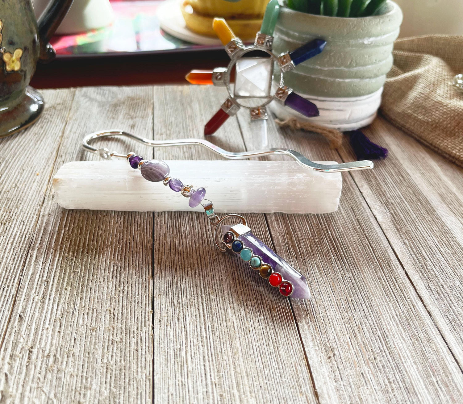 A decorative wand with gemstone beads and a crystal tip rests on selenite atop wood, paired with a crystal chakra decor, potted plant, and the Chakra Amethyst Metal Bookmark—a Zen Gift for Book Lovers.