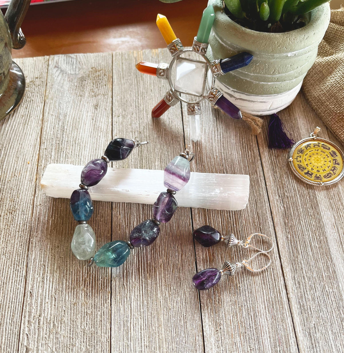 The Fluorite Jewelry Set—Teal and Purple Stone Bracelet and Earrings—rests on a selenite stick atop wood, surrounded by chakra crystals, a potted plant, and a medallion for a vibrant, harmonious display.