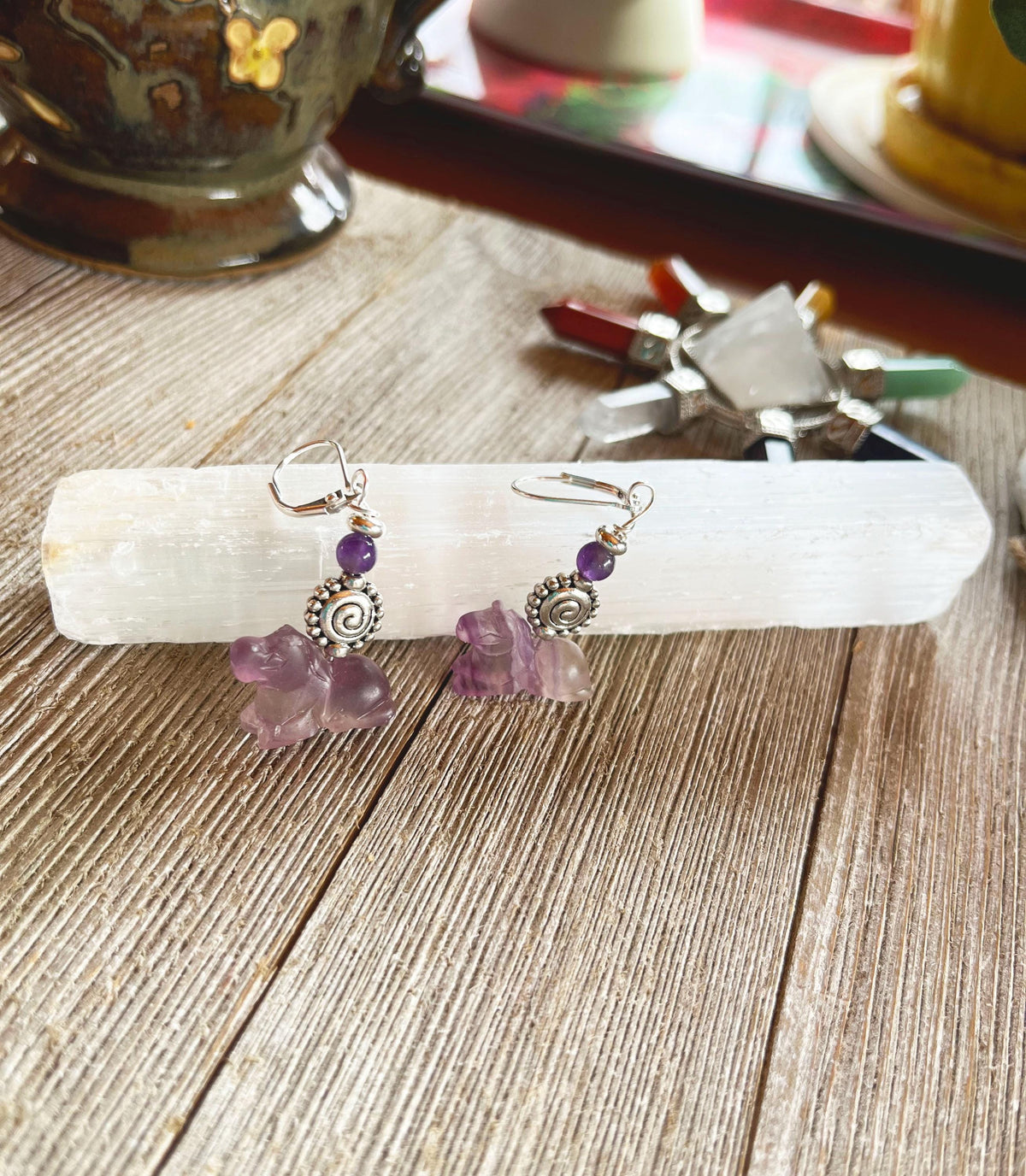 A pair of Horse Earrings Pony Equestrian Jewelry Handcrafted Carved Stone with silver spiral accents and purple stones rest on a white crystal wand atop wood, with colorful crystals and a cup in the background.