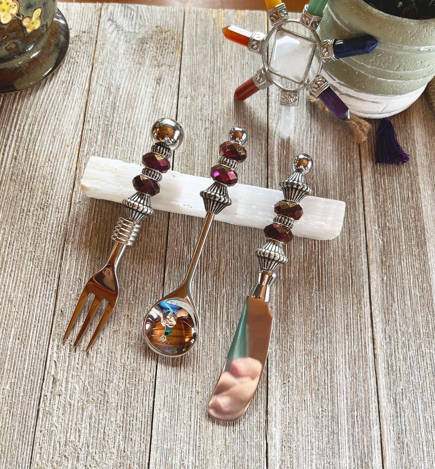 The Purple Beaded Utensil Set—elegant appetizer fork, spoon, and knife—rests on a white crystal holder on wood, complemented by a decorative turtle and potted plant for an elegant entertaining scene.