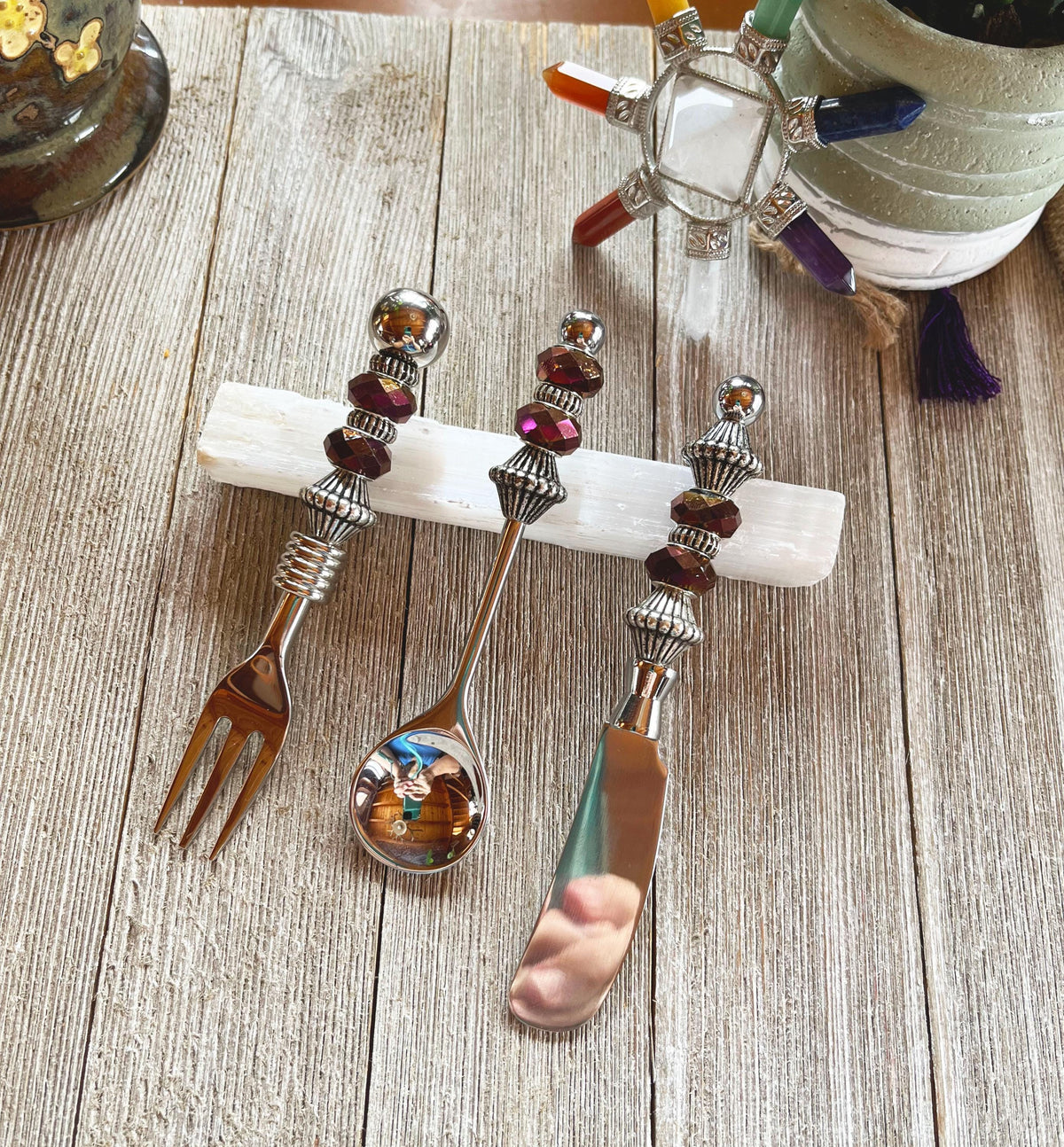 The Purple Beaded Utensil Set—elegant appetizer fork, spoon, and knife—rests on a white crystal holder on wood, complemented by a decorative turtle and potted plant for an elegant entertaining scene.