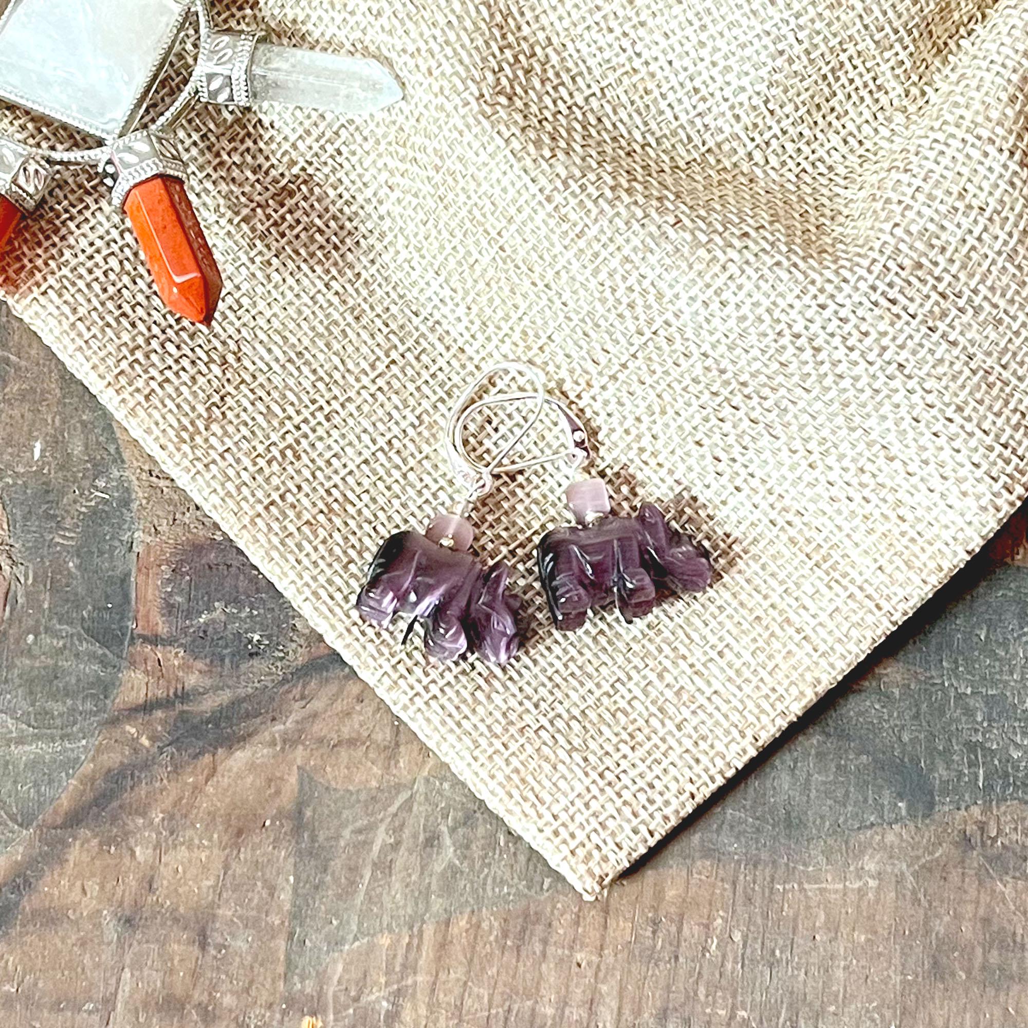 Handcrafted Purple Rhino Earrings with optic stone—perfect wildlife gift—feature purple rhino shapes and silver hooks, displayed on burlap beside a crystal pendant with orange and clear stones atop a rustic wooden surface.