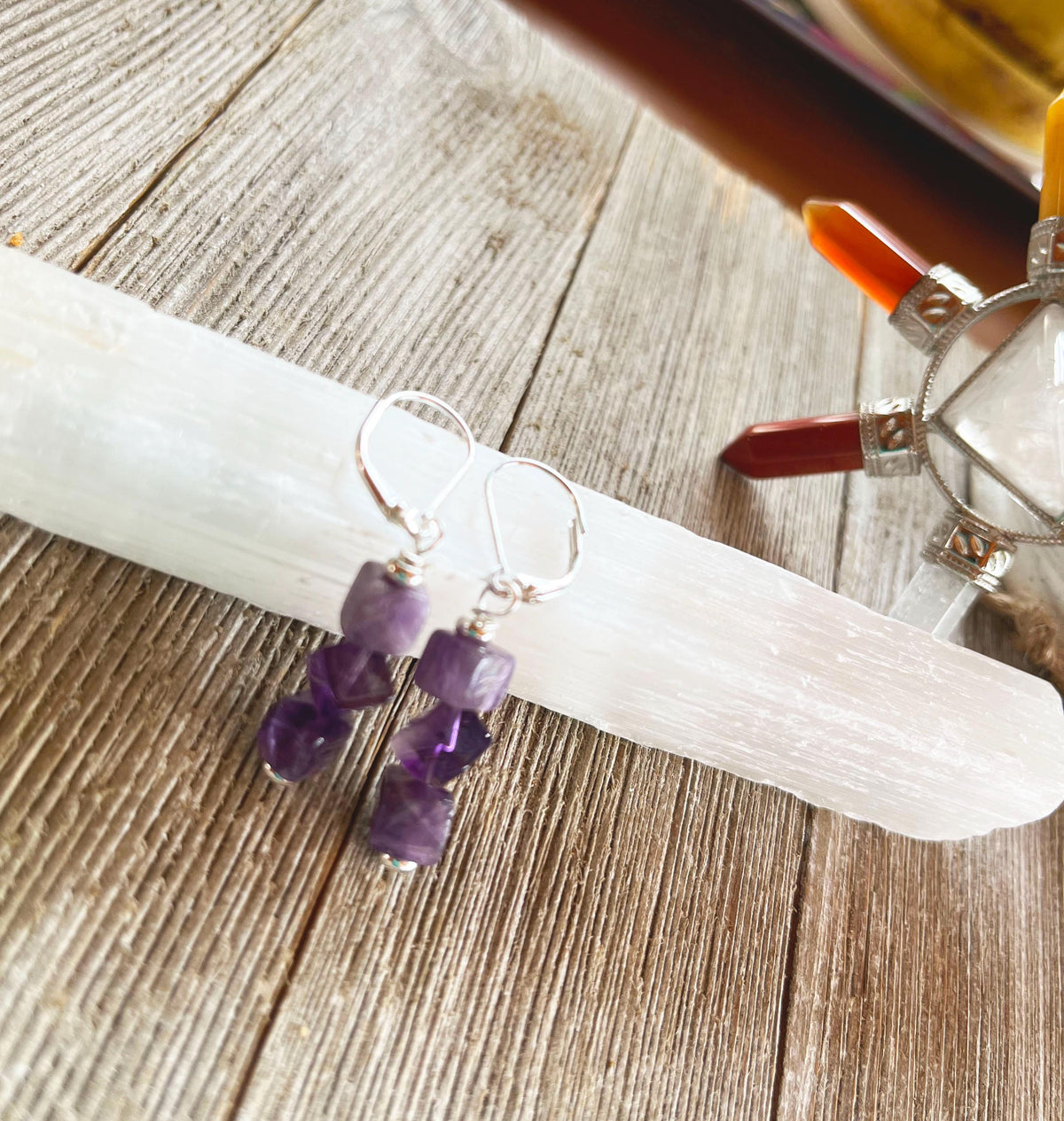 Spiritual Jewelry in Purple Stone Amethyst Cube Earrings—minimalist and handmade in the USA—rest on a white crystal wand atop wood, with a vibrant crystal holder featuring colorful points seen in the background.