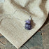 The Serpent Pendant Necklace—handcrafted with wire-wrapped amethyst in a spiritual snake design—rests on textured burlap fabric, with part of a wooden surface visible in the background.