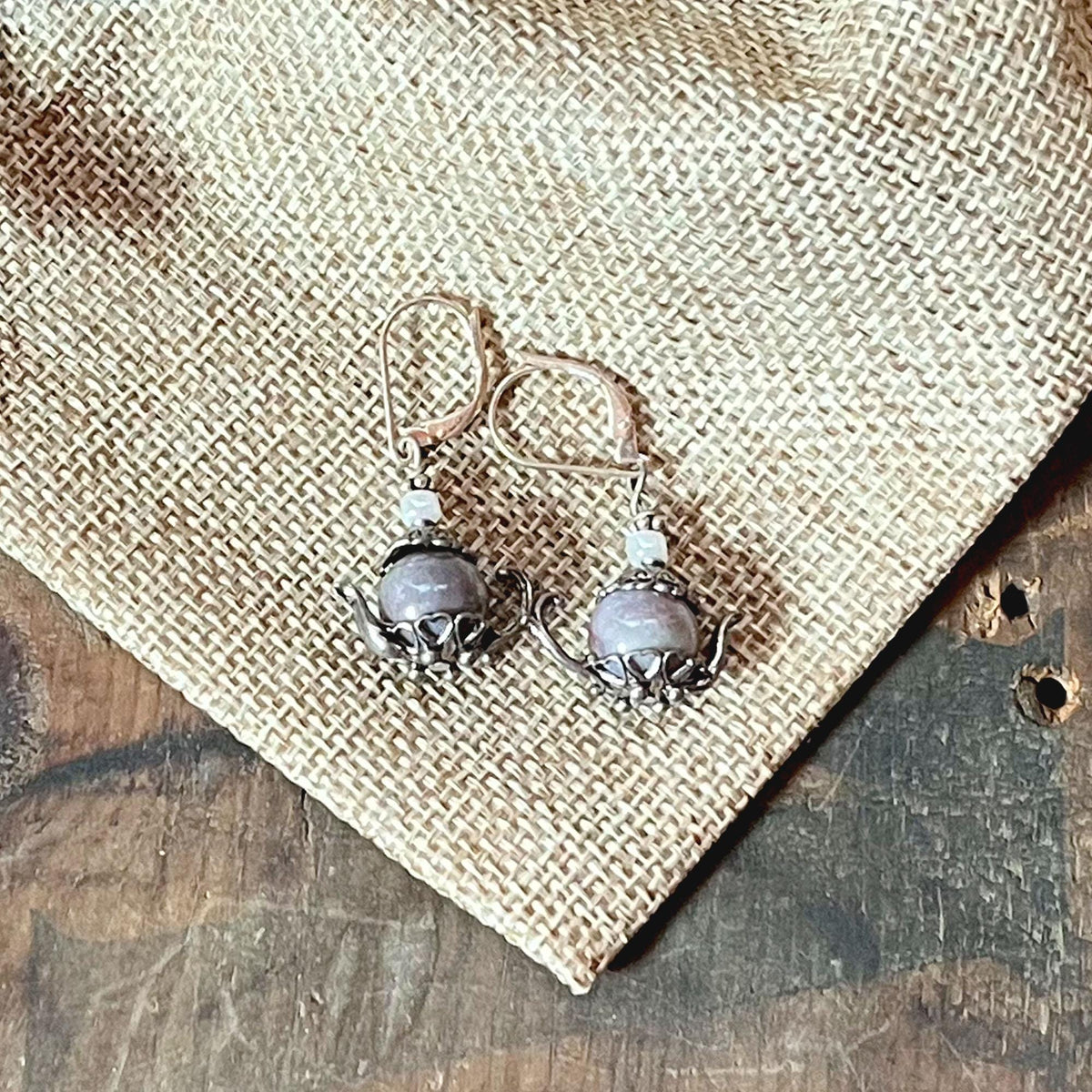 Sterling Silver Teapot Earrings feature round lavender jasper stones and whimsical design, displayed on burlap fabric over a wooden surface.