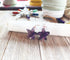 The Purple Amethyst Starfish Earrings: Silver-Plated Nautical Jewelry rest on a white crystal wand atop wood, with colorful crystals blurred in the background—ideal beach jewelry for those who love nautical style.