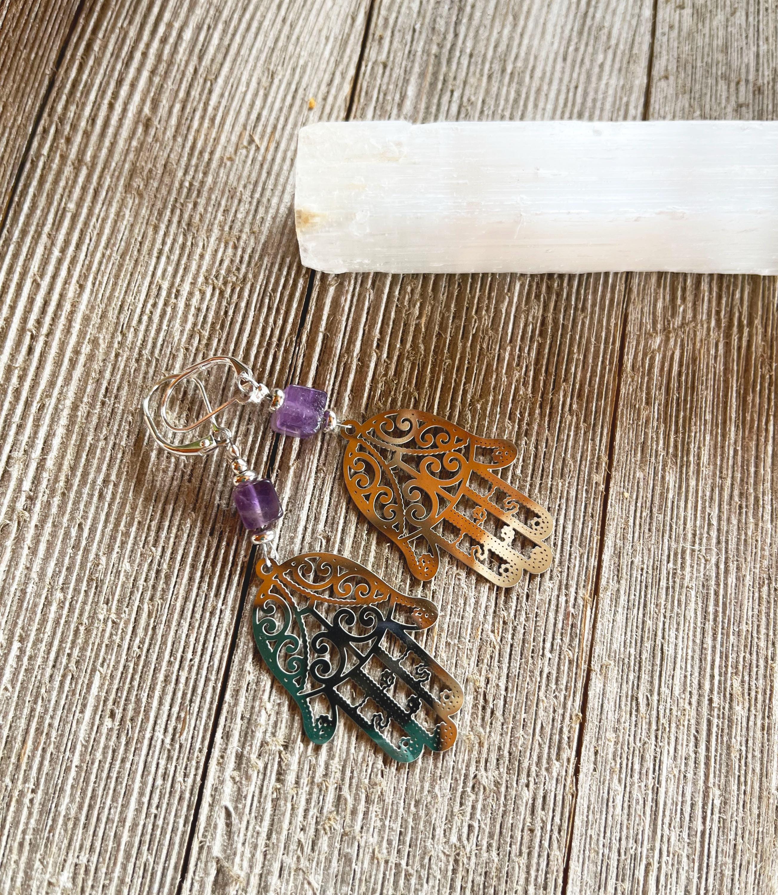 A pair of Purple Earrings Hamsa Hand Charm spiritual jewelry with amethyst cubes rests on a wooden surface beside a white crystal stick.