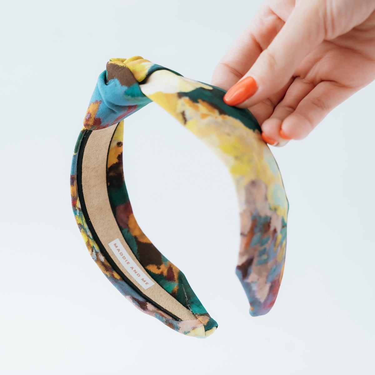 A knotted headband with a colorful abstract print in shades of blue, green, yellow, and brown. The headband is made of fabric and has a black elastic band. The brand name &amp;quot;MADDIE AND ME&amp;quot; is printed on the inside of the headband.