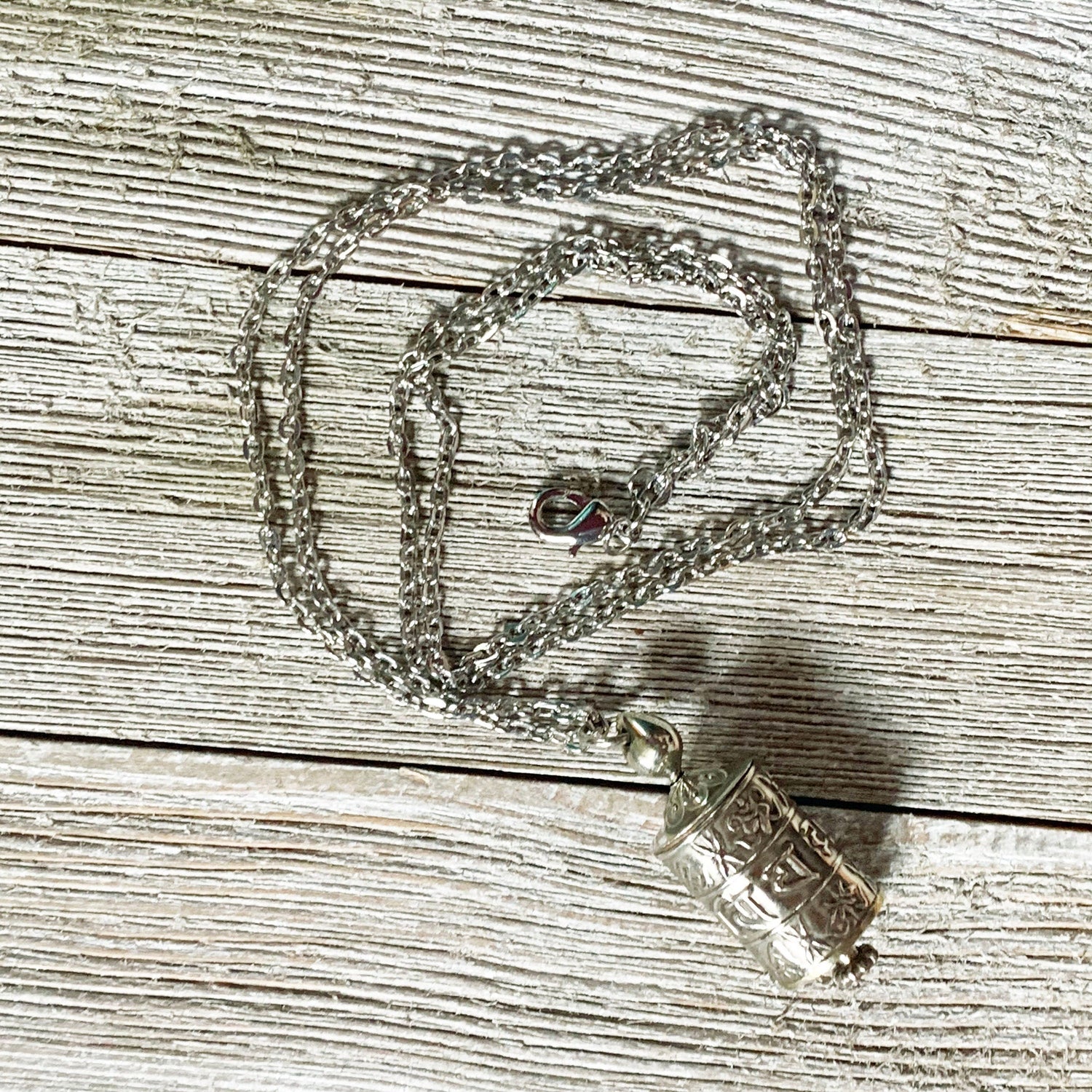 The Prayer Wheel Necklace features a silver Zen Mantra Pendant with intricate patterns, designed for mindful meditation, shown resting on a textured wooden surface.