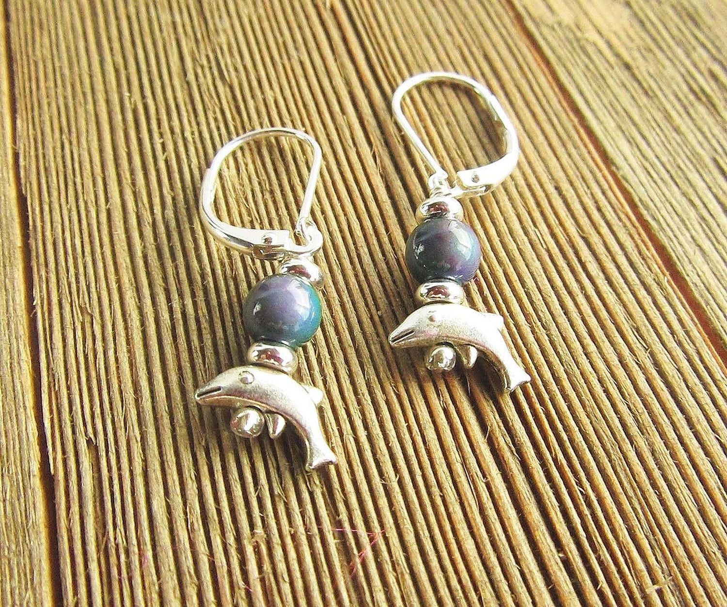 Handcrafted Dolphin Earrings feature small silver dolphin charms and dark color changing mood beads, displayed on a slatted wooden surface—a unique boho ocean jewelry addition.