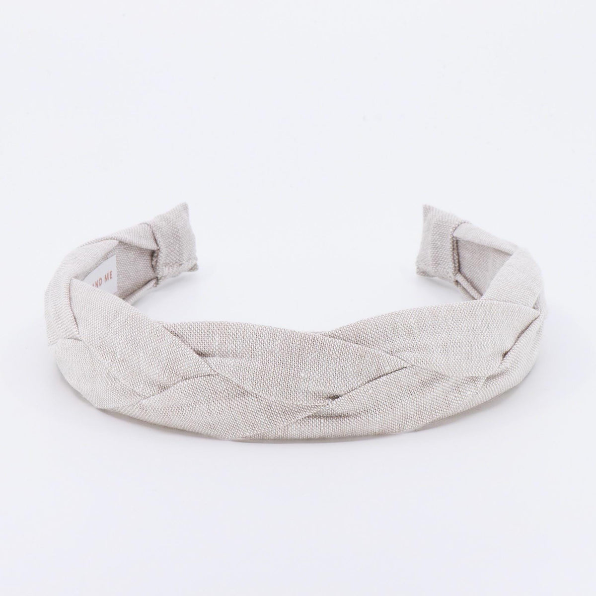 A braided headband in a neutral beige color. The headband is made of a woven fabric and has a thick, textured appearance. The ends of the headband are folded over and sewn. The headband is designed to be worn on the head.
