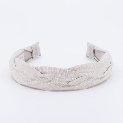 A braided headband in a neutral beige color. The headband is made of a woven fabric and has a thick, textured appearance. The ends of the headband are folded over and sewn. The headband is designed to be worn on the head.