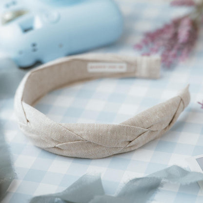 A beige braided headband with a textured appearance. The headband is made of fabric and has a label on the inside. The background includes a light blue object and a light blue and white checkered surface.