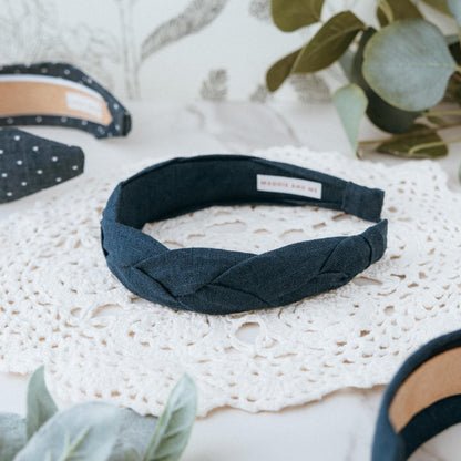 A navy blue braided headband with the text &amp;quot;MADDIE AND ME&amp;quot; on a white label. The headband is made of a linen fabric and is displayed on a white lace doily. Other headbands with white polka dots are visible.