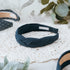 A navy blue braided headband with the text "MADDIE AND ME" on a white label. The headband is made of a linen fabric and is displayed on a white lace doily. Other headbands with white polka dots are visible.