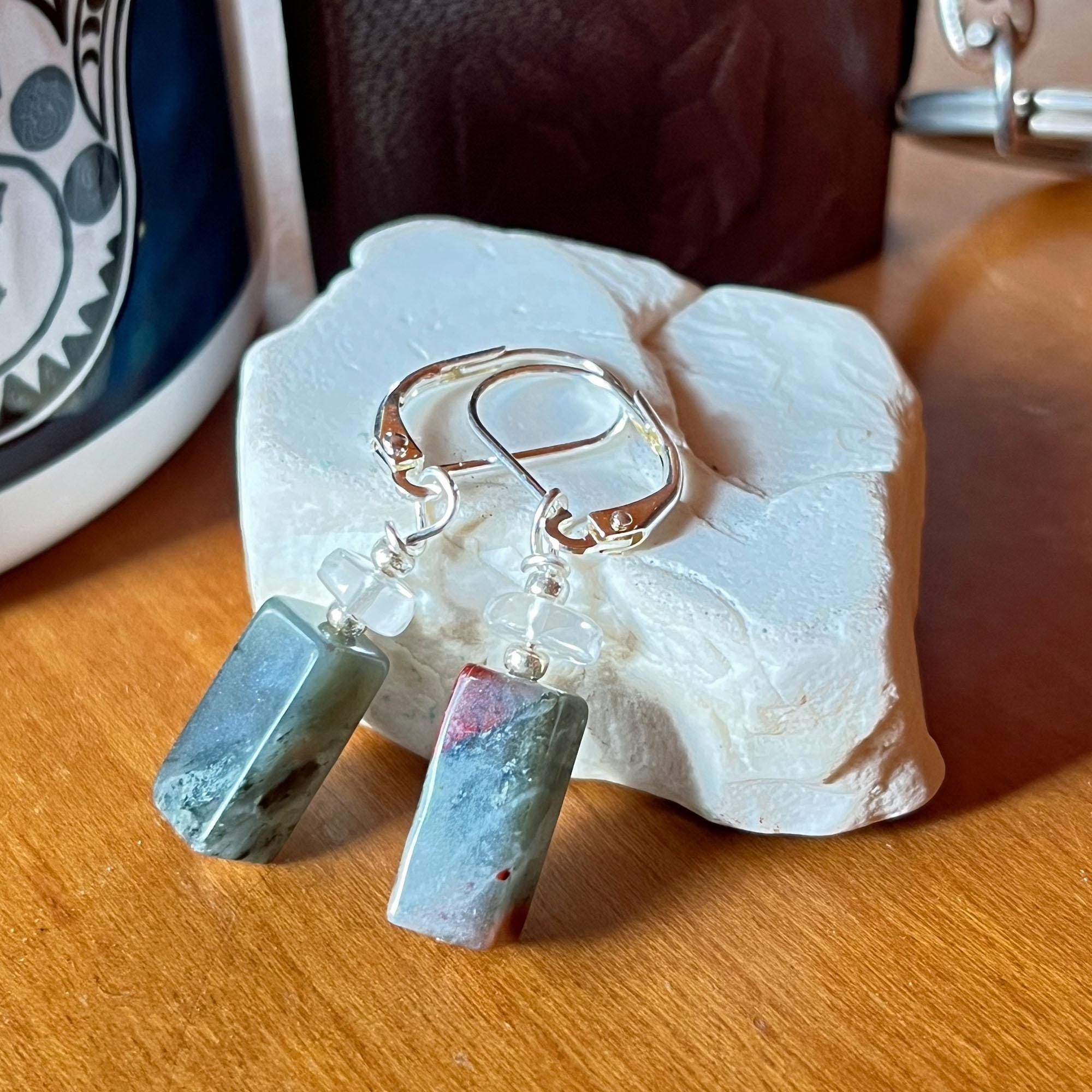 Spiritual Bloodstone Dangle Earrings with Clear Quartz: minimalist Zen jewelry featuring silver hoops, rectangular green and gray stone pendants, and translucent beads, displayed on white textured stone near a ceramic cup.