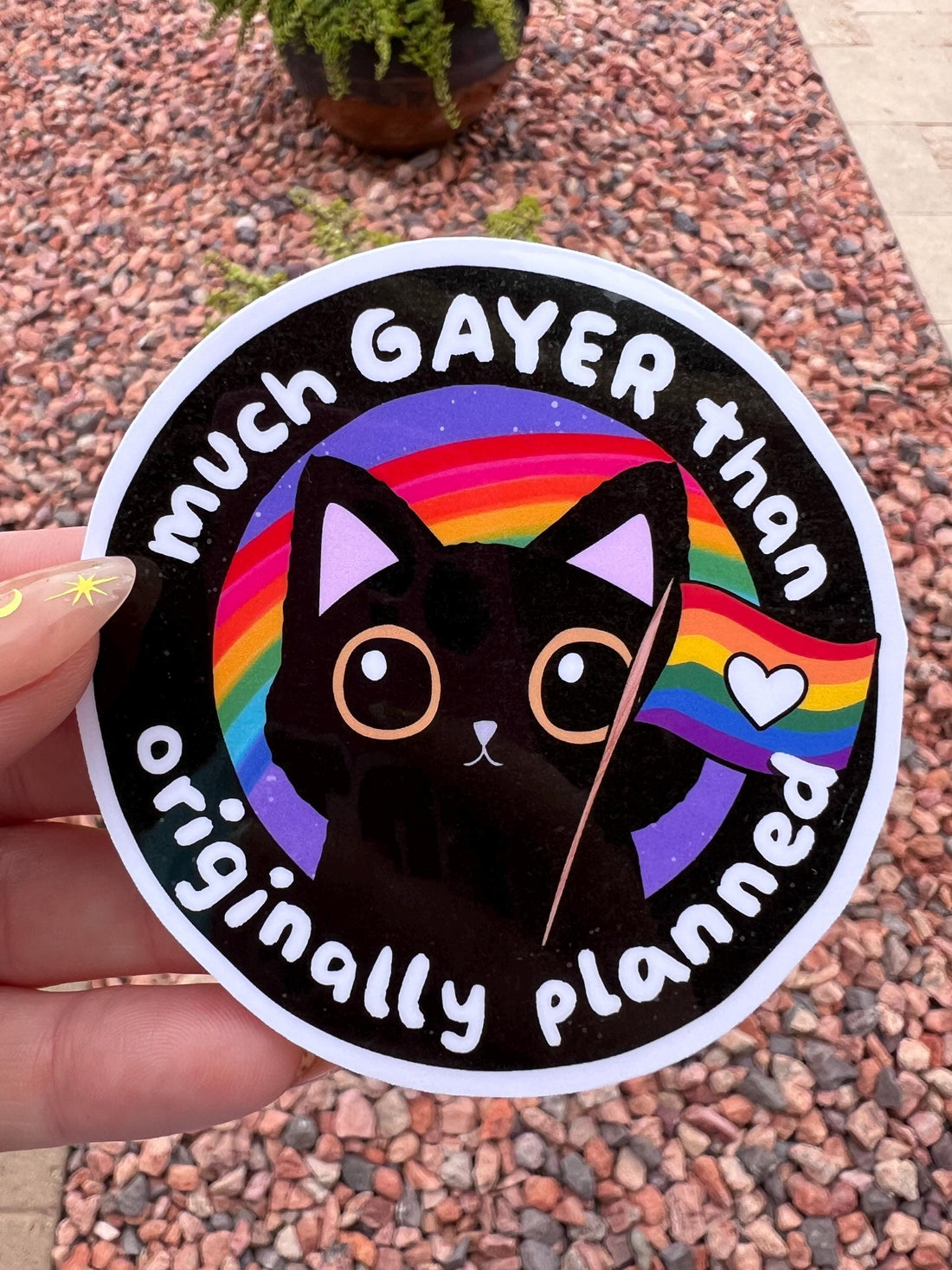 May include: A round sticker with a black outline and a black cat illustration. The cat is holding a rainbow flag with a white heart. The sticker has a rainbow background and the text &amp;
