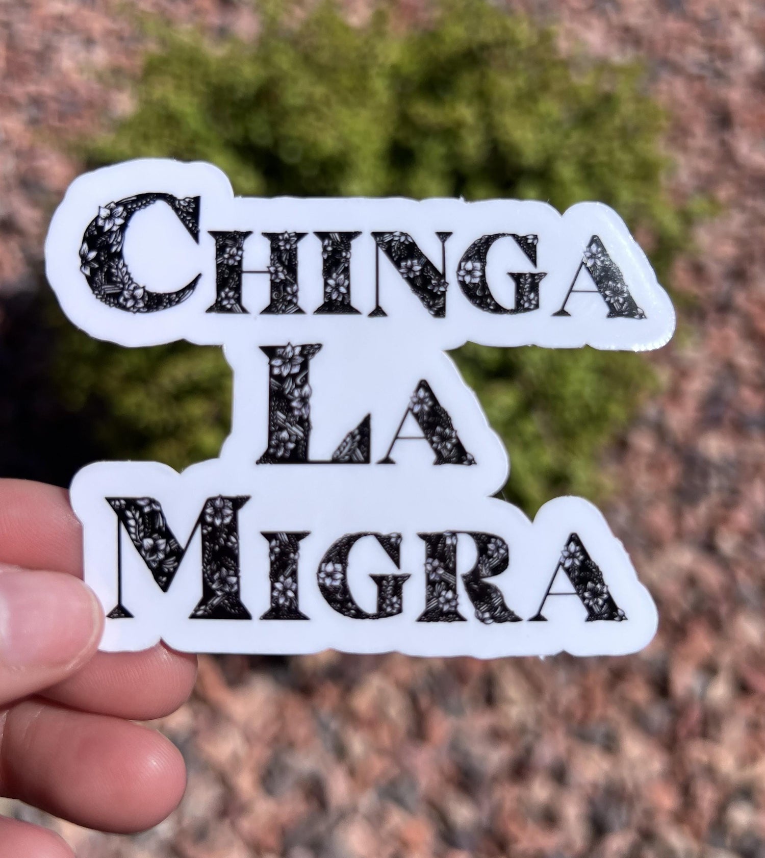 May include: A white sticker with the words &amp;quot;CHINGA LA MIGRA&amp;quot; in black, floral-patterned letters.