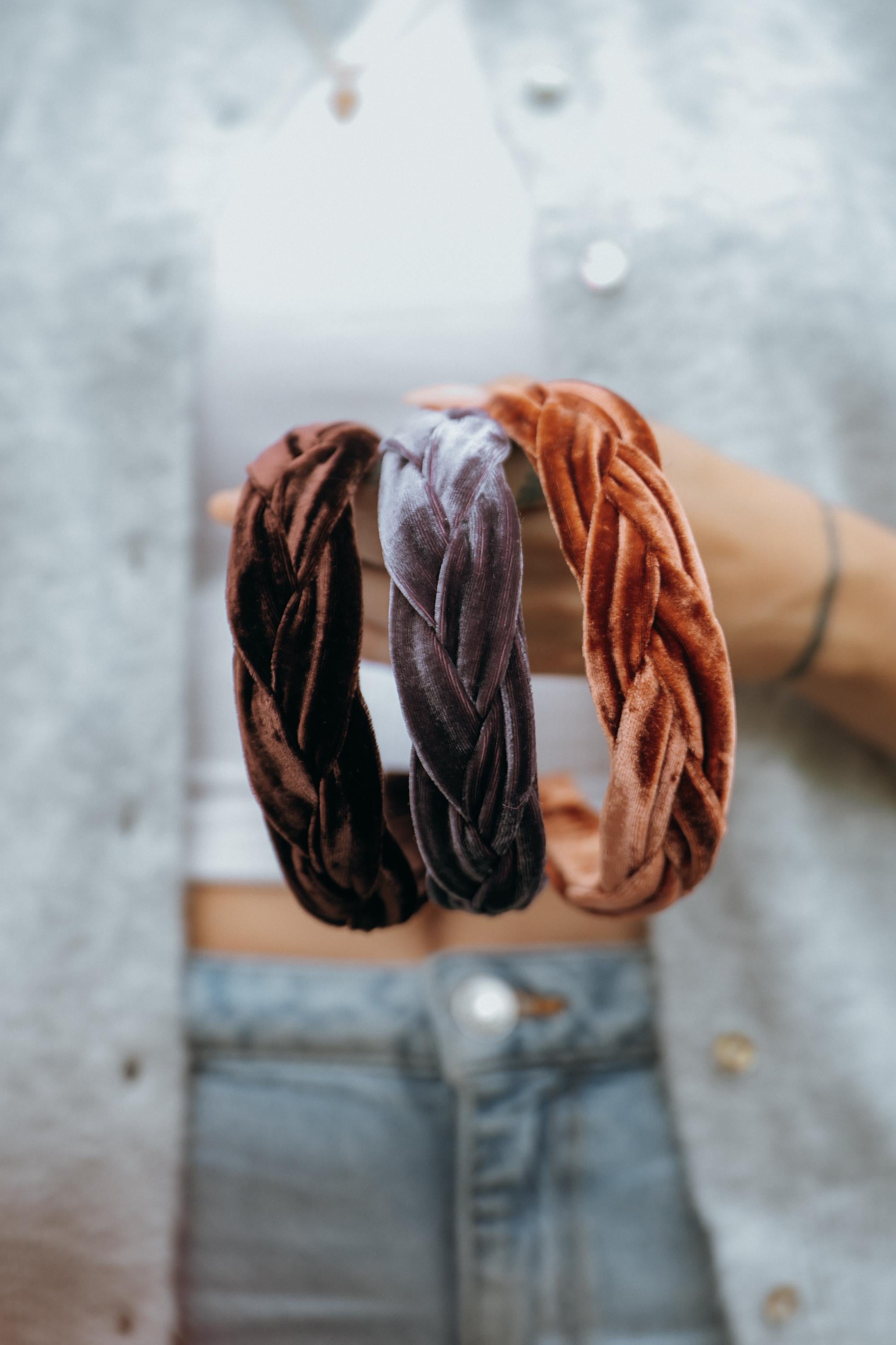 May include: Three braided velvet headbands in shades of brown, mauve, and orange. The headbands are held up against a person&amp;