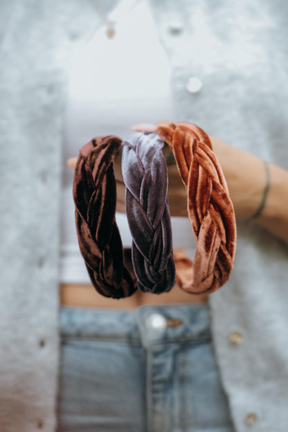May include: Three braided velvet headbands in shades of brown, mauve, and orange. The headbands are held up against a person&amp;
