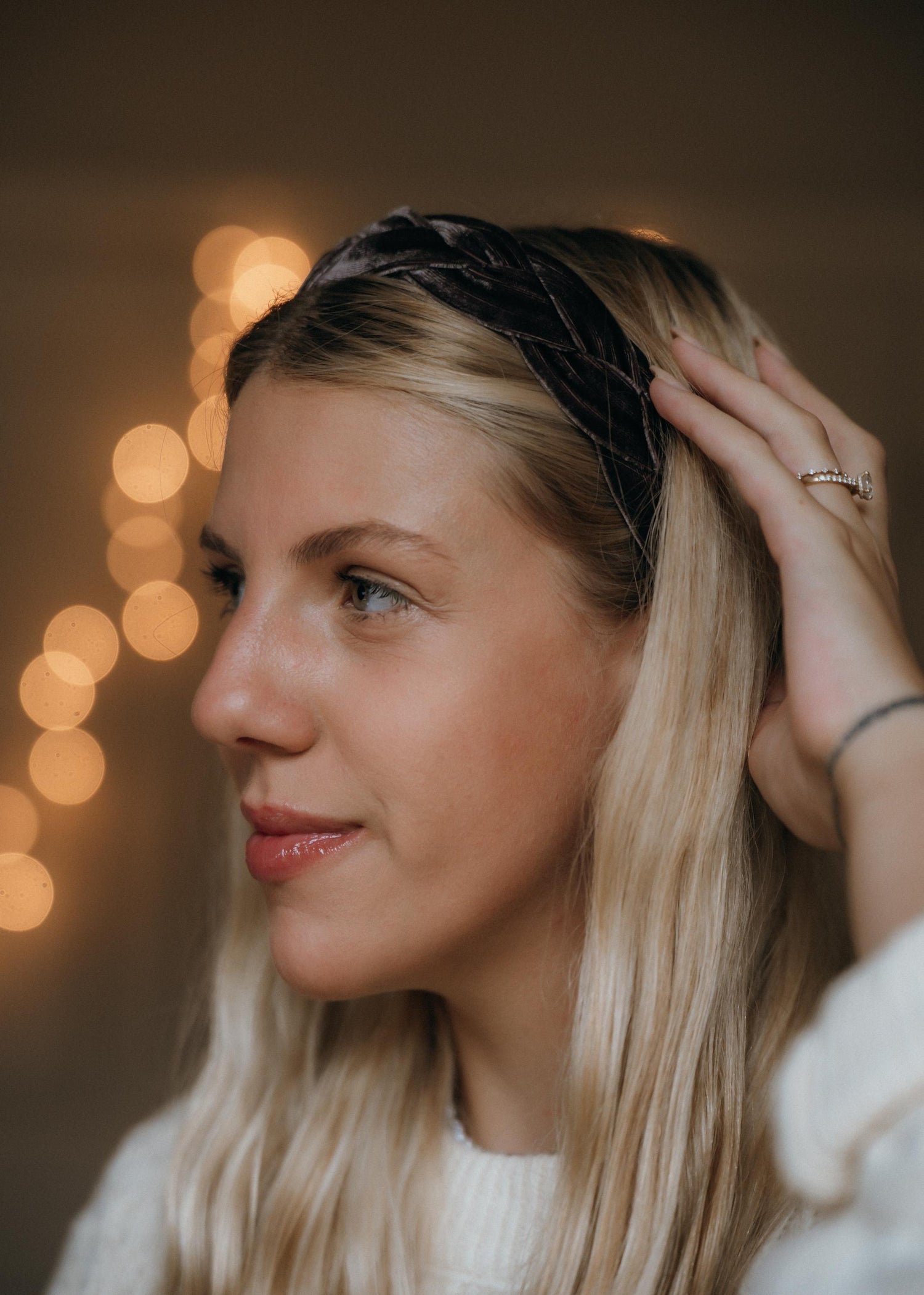 May include: A side profile of a person with blonde hair wearing a mauve braided velvet headband. The person is wearing a white sweater and a ring on their finger. The background has soft, blurred lights.