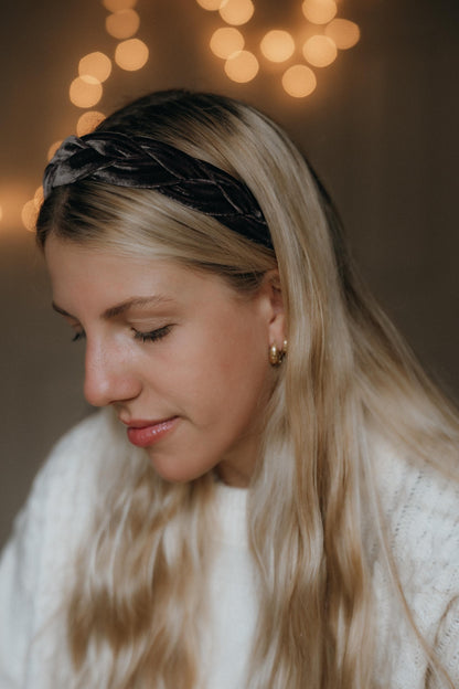 May include: A mauve velvet braided headband. The headband is worn by a person with long blonde hair and gold hoop earrings. The person is wearing a white sweater. The background has soft, out-of-focus lights.