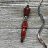 The Handcrafted Red Jasper Goddess Bookmark features a wavy silver handle, a red jasper bead, and a red goddess charm on metal loops—a unique Divine Feminine Gift displayed on a textured wooden surface.