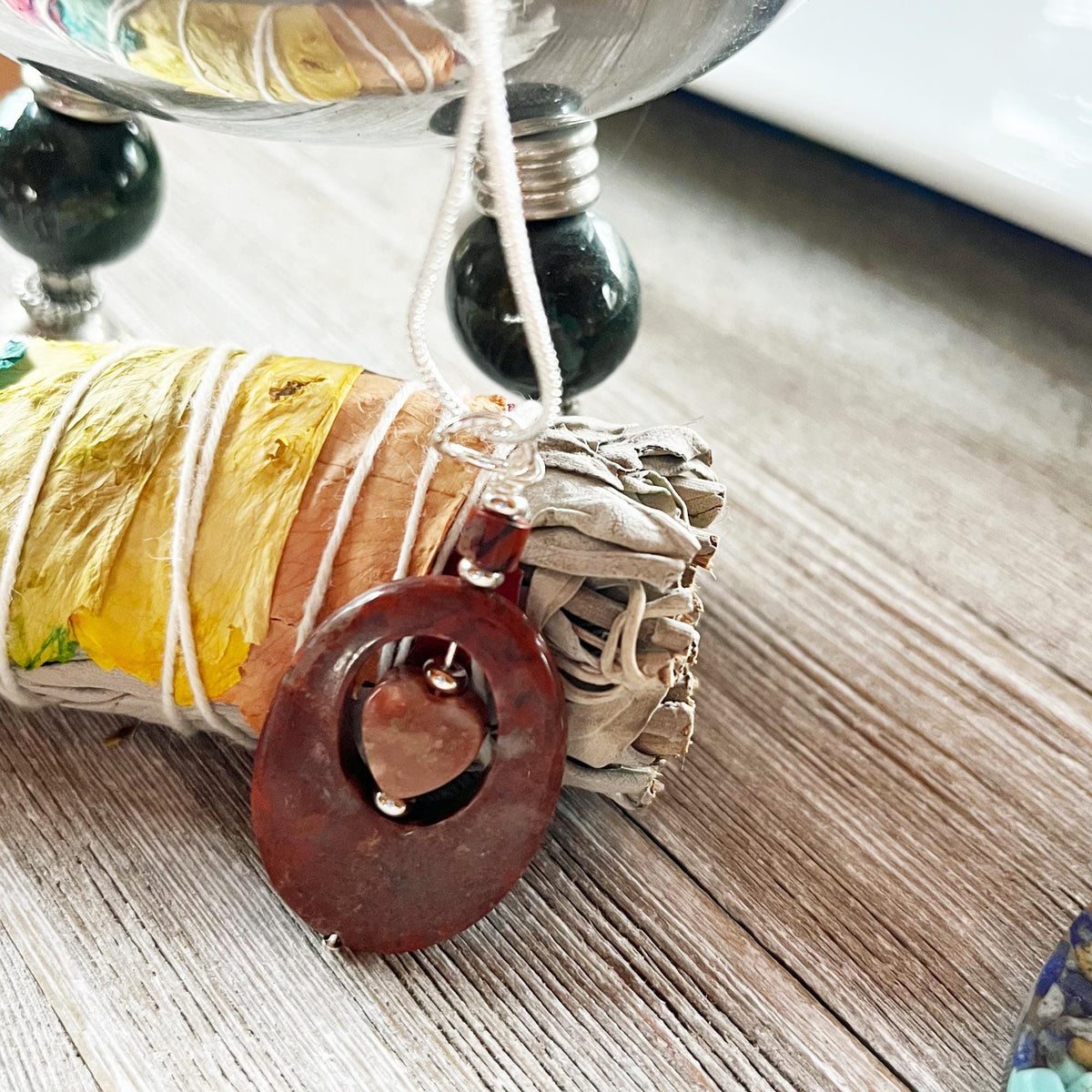 The Red Jasper with a Heart Spinner Pendant necklace features a circular jasper stone on a white cord, displayed atop dried sage and flowers. A silver bowl and decor in the background complement this grounding fidget jewelry piece.